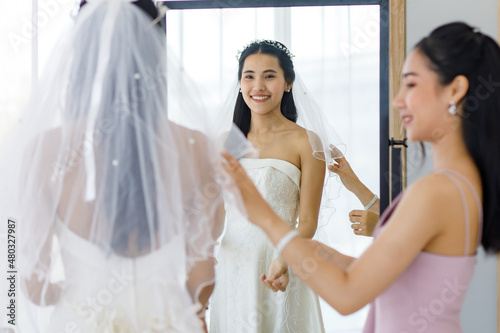 Asian lovely beautiful happy cheerful female bride in white long wedding dress standing staring at her reflection in mirror while pretty bridesmaid helping adjusting facial veil in blurred foreground