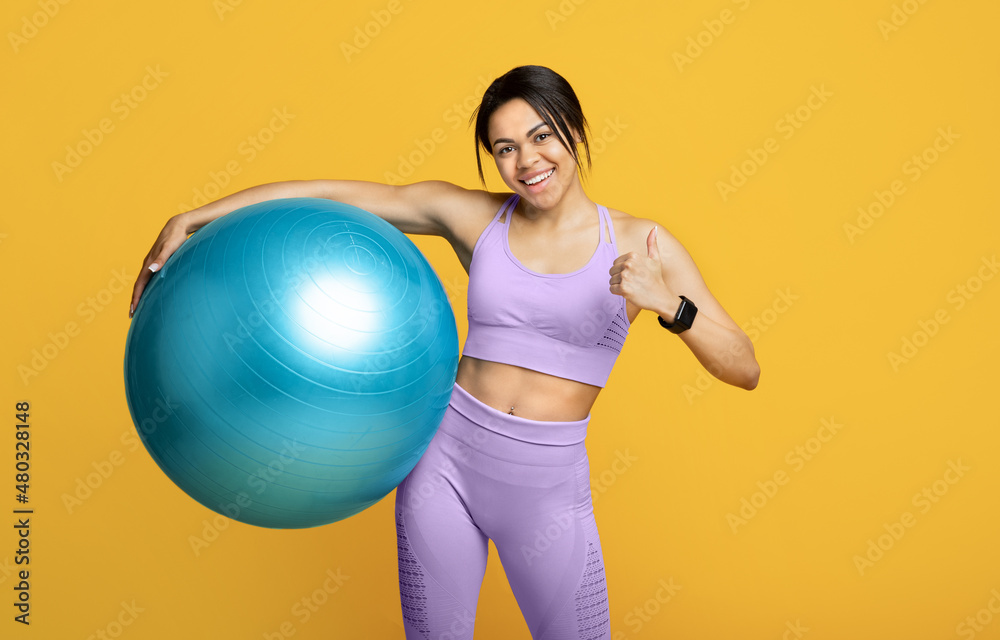 Young sporty black woman holding fitness ball and showing thumb up, standing on yellow background Young sporty black woman holding fitness ball and showing thumb up, standing on yellow background