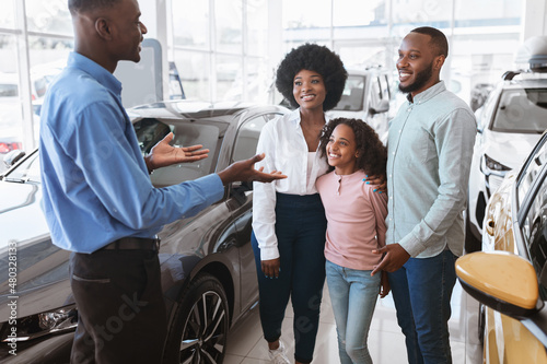 Car selling business. Confident manager showing luxury automobile to black family at auto dealership center