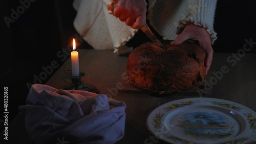 A woman cutting a panettone a Italian Christmas cake lying on a board on a wooden table near a knife, a napkin, a plate and a burning candle.