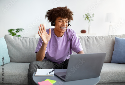 Happy black teenager in headphones greeting his friend or teacher online, waving at camera from home