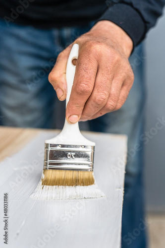 Close up on white paint brush in the hand of unknown man painting wooden board hobby and renovation repairing concept