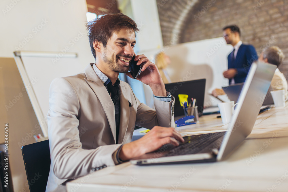 Happy businessman using laptop and phone with team discussing project in the background. Happy businessman using laptop and phone with team discussing project in the background.