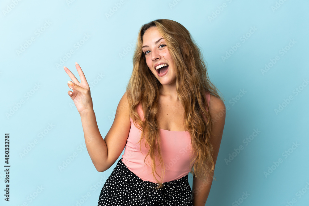 Young blonde woman isolated on blue background smiling and showing victory sign Young blonde woman isolated on blue background smiling and showing victory sign