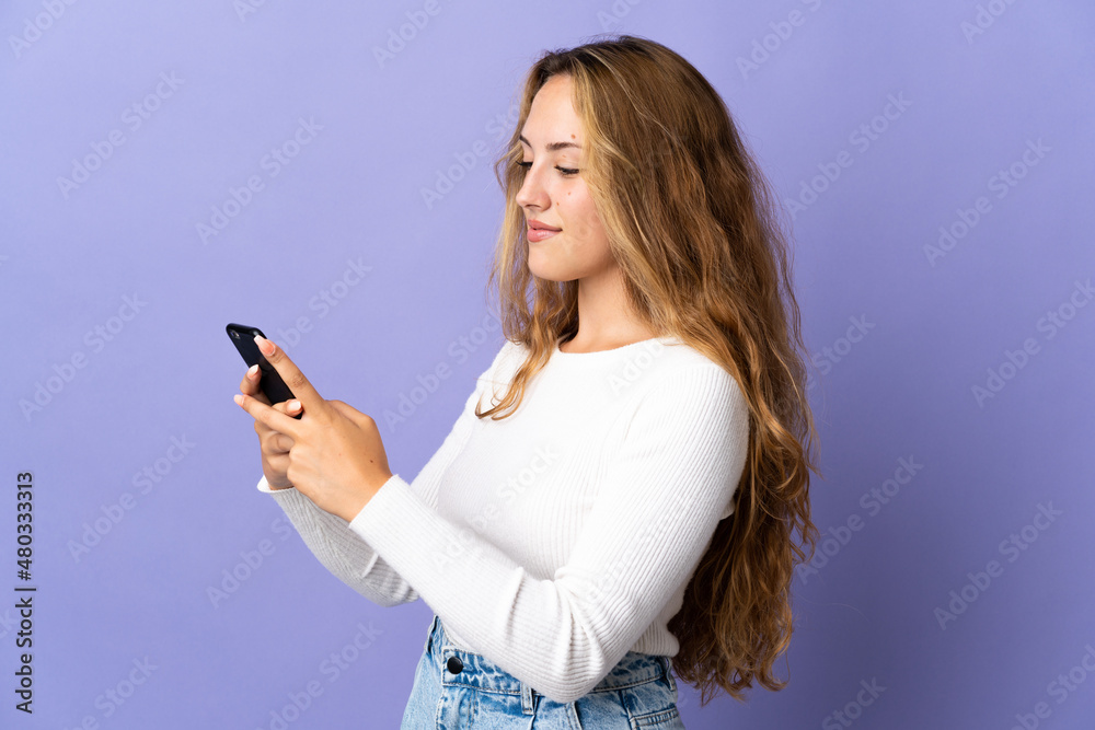 Young blonde woman isolated on purple background sending a message or email with the mobile Young blonde woman isolated on purple background sending a message or email with the mobile