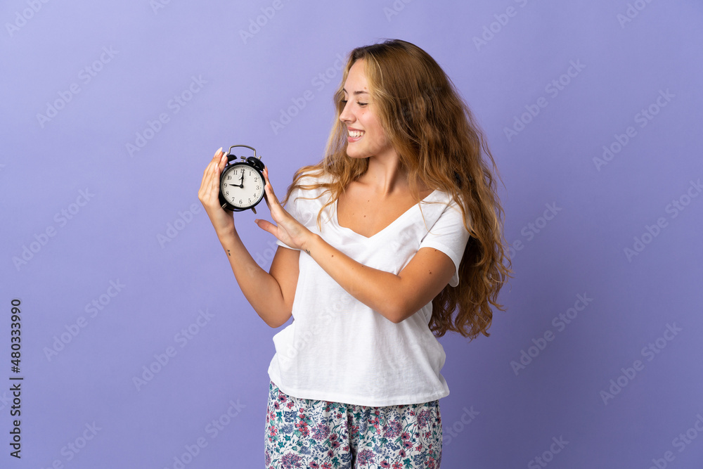 Young blonde woman isolated on purple background in pajamas and holding clock with happy expression Young blonde woman isolated on purple background in pajamas and holding clock with happy expression