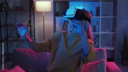 Female gamer. Virtual reality. New experience. Amazed woman in vr goggles showing surprise playing cyber game sitting sofa dark neon light home interior.