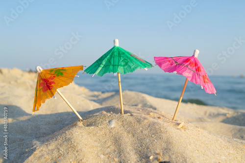 Three small paper cocktail umbrellas stand in sand on sandy beach close-up. Small paper umbrellas on sandy shore near sea and sea waves on sunny summer day. Leisure, Vacation, Travel, Tourism Concept © mari1408