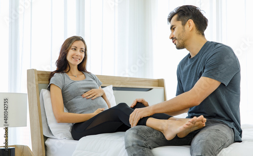 Lovely husband do prenatal massage on legs of beloved pregnant wife with maternity care on bed to release stress and muscle pain while mom tenderly embrace belly of expecting unborn baby.