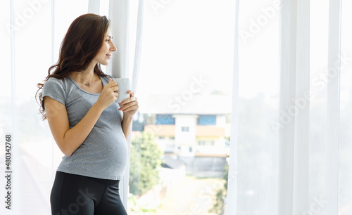 Happy Caucasian woman drinks glass of healthy beverage for belly nourishment of pregnancy and calmly relax by looking out window