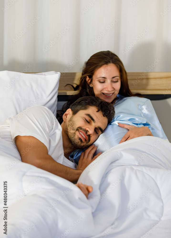 Fototapeta premium Happy Caucasian adult couple laugh and smile while talking fun and embrace each together on bed as enjoy resting under warm quilt to sleep in bedroom on romantic night at home