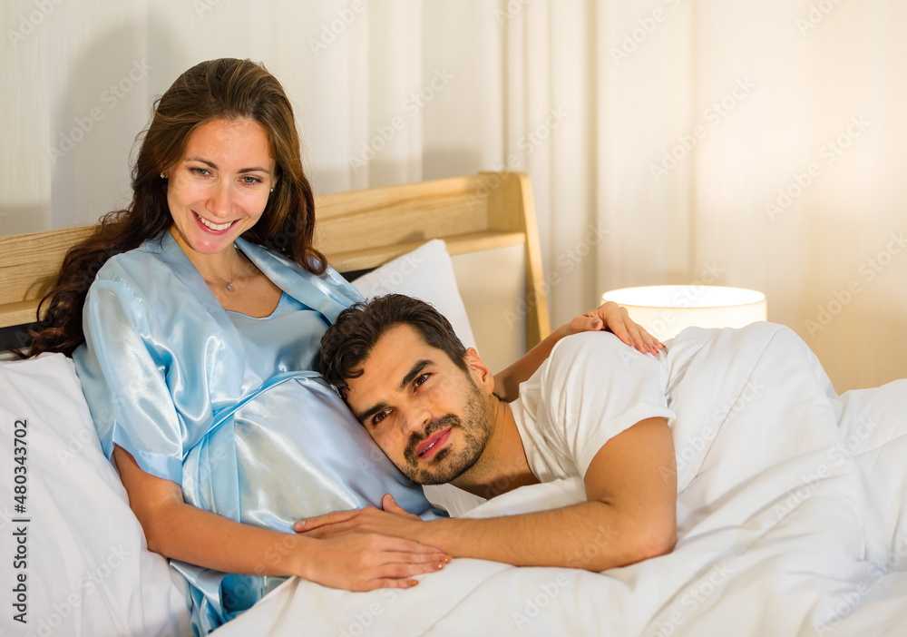 Fototapeta premium Happy Caucasian husband tenderly lies down on belly and embrace lovely pregnant wife who smile on bed
