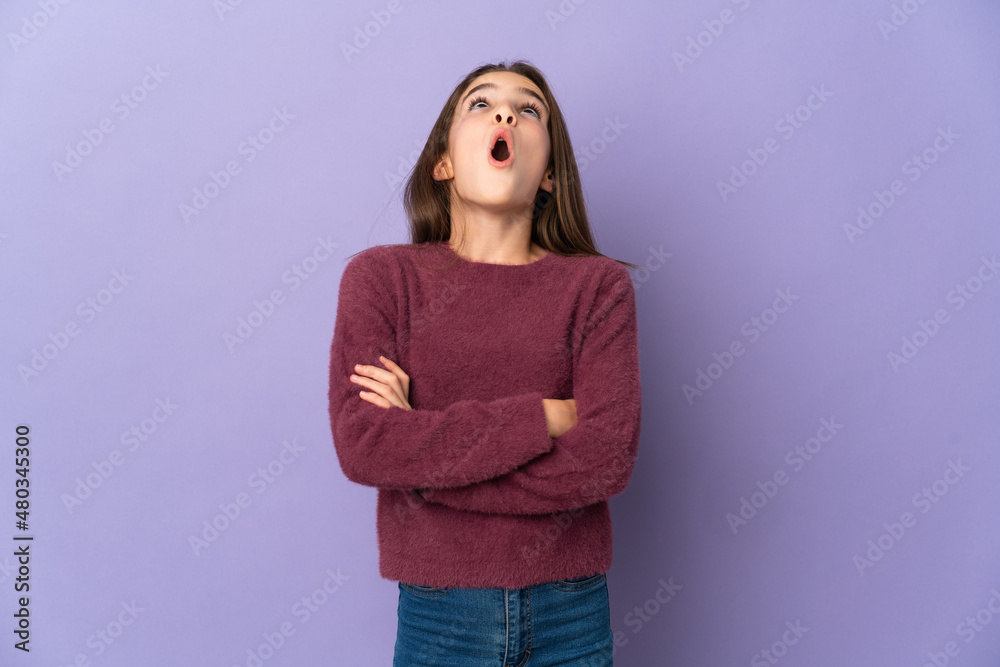 Little girl isolated on purple background looking up and with surprised expression