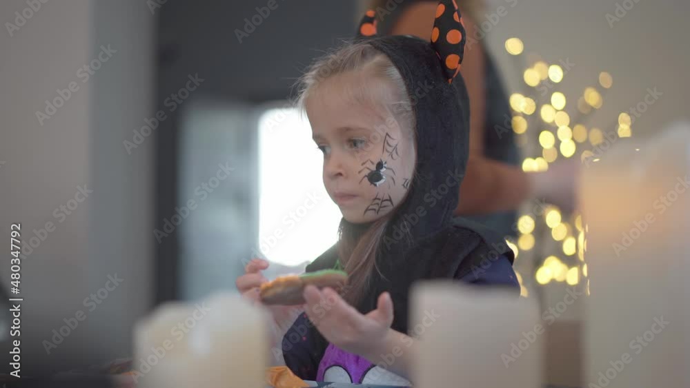 Little girl eat icing just making handmade Halloween cookies sitting on the table dressed Halloween carnival costume with horns . Halloween party in big family. High quality 4k footage