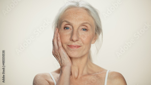 Closeup Portrait of Senior Woman Looking at Camera  Touching Hair  Beautiful Face  Smiling. Elderly Lady with Natural Grey Hair  Blue Eyes. Abstract Beige Background  Tender and Soft Moment