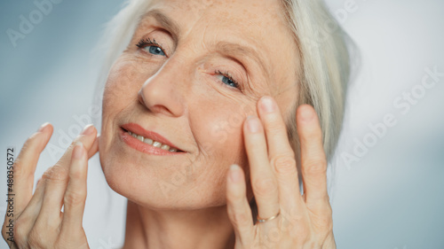 Portrait of Senior Woman Looking at Camera, Touching Beautiful Face, Smiling. Elderly Female with Natural Grey Hair, Blue Eyes, Soft Skin. Dignified Old Age Concept for Skincare and Healthy Living © Gorodenkoff