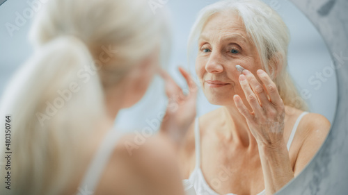 Portrait of Beautiful Senior Woman Morning Routine, Looking into Mirror Gently Applying Face Cream. Elderly Lady Makes Her Skin Soft, Smooth, Wrinkle Free with Natural anti-aging Cosmetics, Products