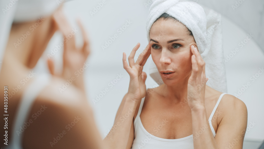 Beautiful Caucasian Woman Touches Her Perfect Face, Enjoying Her Ideal Wrinkle Free Skin, Sensually Smiles into Bathroom Mirror. Happy Middle Aged Female Uses Natural Cosmetic Skincare Products