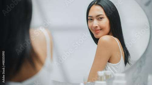 Beautiful Asian Woman Touches Her Perfect Soft Shoulder, Neck, Sensually Smiles in the Mirror. Happy Female Enjoying Her Beauty Routine. Wellness Natural Cosmetic Skincare Products.