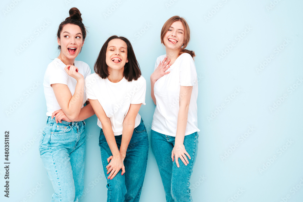 Obraz premium Three young beautiful smiling hipster female in trendy same summer white t-shirt and jeans clothes. Sexy carefree women posing near light blue wall in studio. Cheerful and positive models having fun