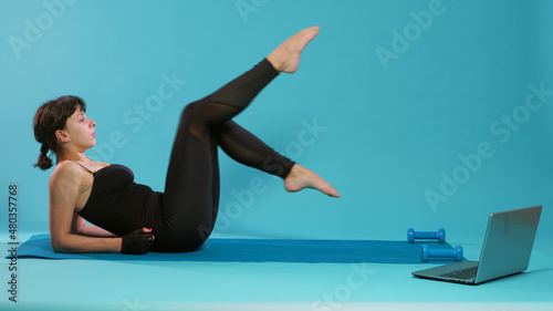 Fit woman training legs muscles with bicycle exercise over blue background. Adult exercising in front of camera, doing physical activity and looking at training video to practice sport.
