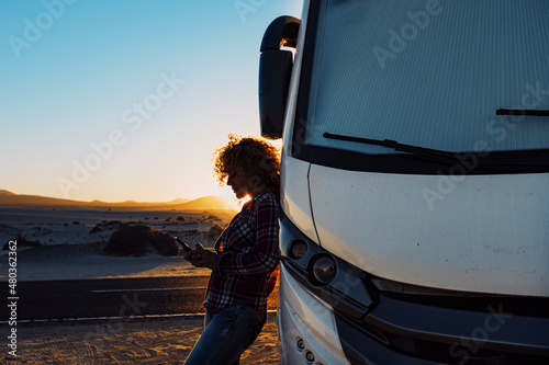 People on travel with modern camper. Woman using mobile phone roaming against big motorhome. Concept of modern lifestyle and travelers. Vacation and vanlife sending message with cellular.
