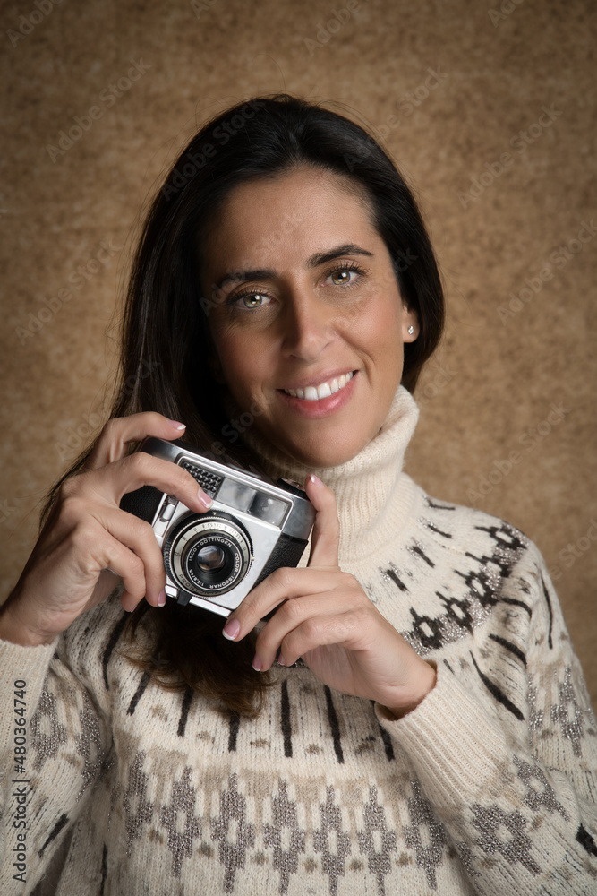 Retrato de joven mujer atractiva sonriente con cámara de fotos retro, con fondo desenfocado marrón y jersey blanco.