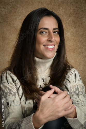 Retrato de joven mujer atractiva sonriente con las manos juntas , con fondo desenfocado marrón y jersey blanco.