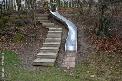 stainless steel slide on the playground with concrete hills and rocks and wooden staircase. Large glossy tube for sliding children with an impact on the tartan rubber pad. the tube is an open trough