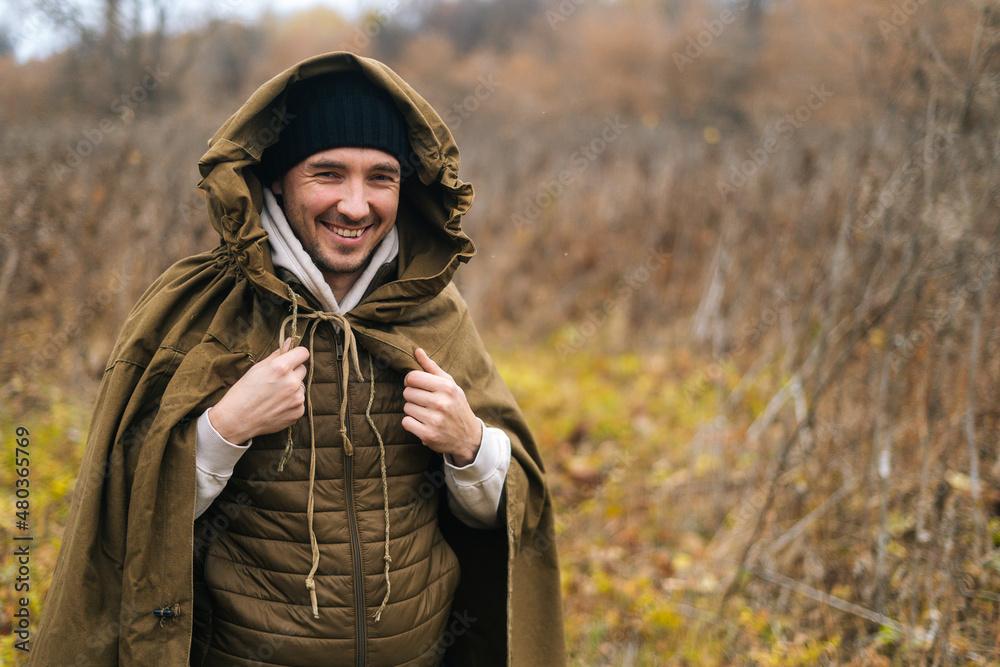 Portrait of cheerful hiker man wearing green raincoat tent standing in thicket of bushes in cold overcast day and looking at camera. Concept of scout, research, travel and survival in nature.