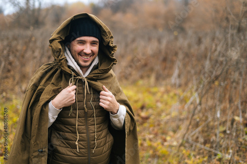 Portrait of cheerful hiker man wearing green raincoat tent standing in thicket of bushes in cold overcast day and looking at camera. Concept of scout, research, travel and survival in nature.