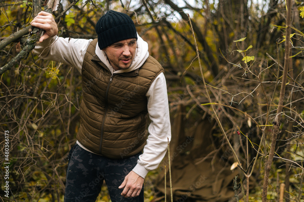 Portrait of exhausted lost tourist male wearing warm clothes wading through thickets in forest in cold overcast day. Concept of scout, research, travel and survival in nature.