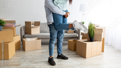 Moving into own house. Loving black husband lifting his wife and having fun among boxes in new apartment, crop