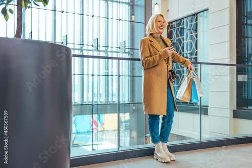 Young beautiful girl is shopping in the Mall. Woman enjoying the day in the shopping mall. Happy girl with shopping bags texting on smartphone. Shopping concept.