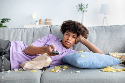Lazy black teen guy lying on couch with scattered snacks, holding remote control, watching TV at home photo