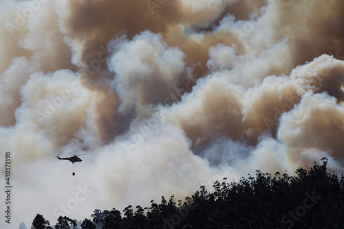 A fire helicopter with a bambi bucket in the clouds of a forest fire