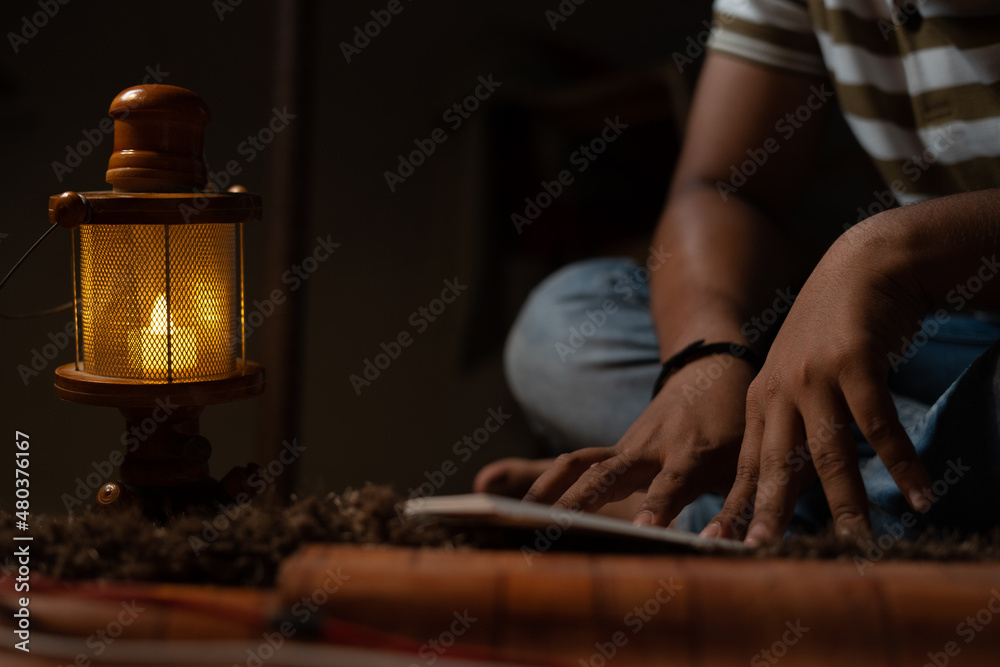 unrecognizable Young student busy reading during night under oil lamp or lantern due to power loss and Poverty - concpet of power cut, blackout, electricity failure during examination. - obrazy, fototapety, plakaty 