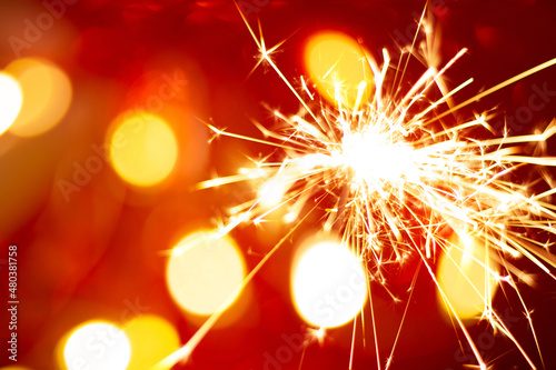 Glowing burning sparkler with gold blurred bokeh on red background. Concept holidays of Christmas and Happy New Year or Birthday. Hand-held firework