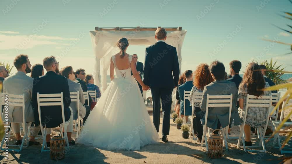 Beautiful Bride in White Wedding Dress and Handsome Groom in a Traditional Black Suit Going Down the Aisle at an Outdoors Ceremony Venue Near the Sea with Happy Multiethnic and Diverse Friends.
