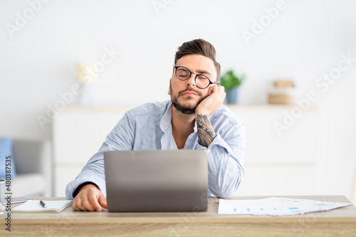 Overworked young freelancer man sitting at workplace and sleeping, napping while working on laptop, copy space
