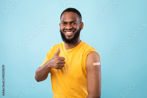 Cheerful Afro man showing thumb up gesture, demonstrating arm with band aid after injection of coronavirus vaccine