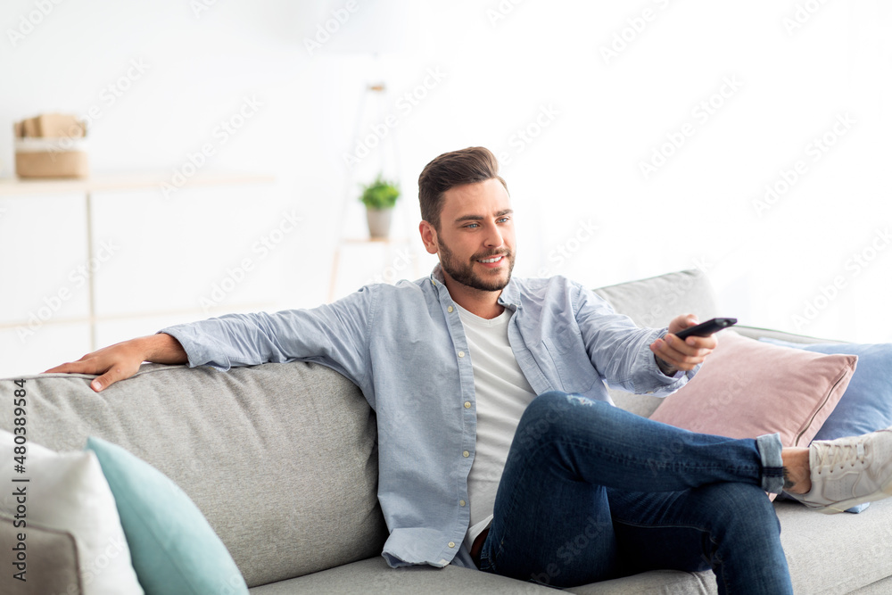 Leisure and entertainment. Happy guy watching TV switching television channels with remote control, sitting on sofa
