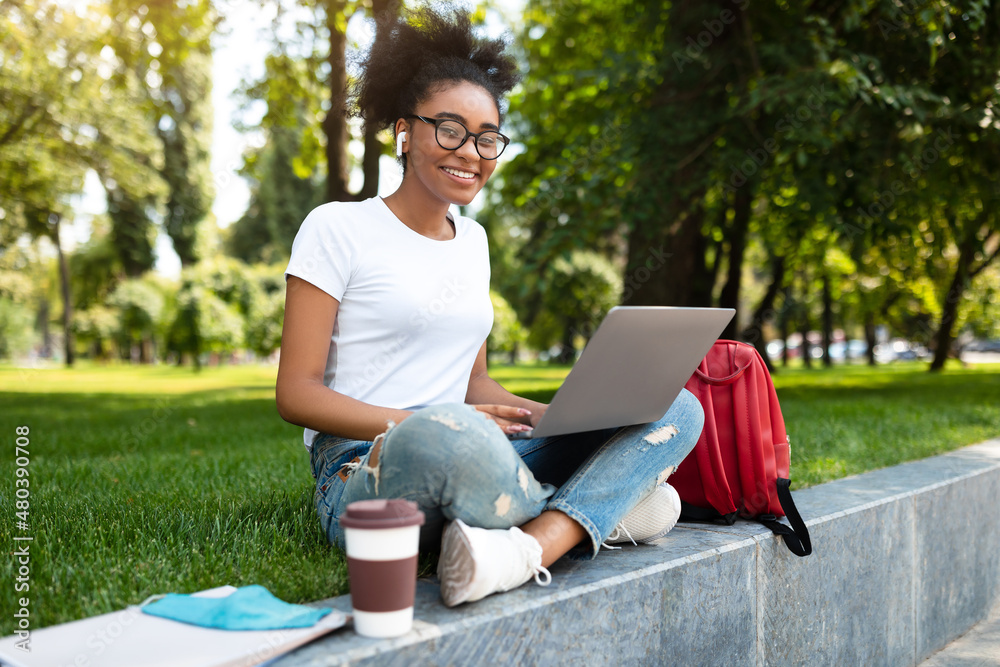 Happy Black Female Student Using Laptop Wearing Earbuds Sitting Outdoors Happy Black Female Student Using Laptop Wearing Earbuds Sitting Outdoors