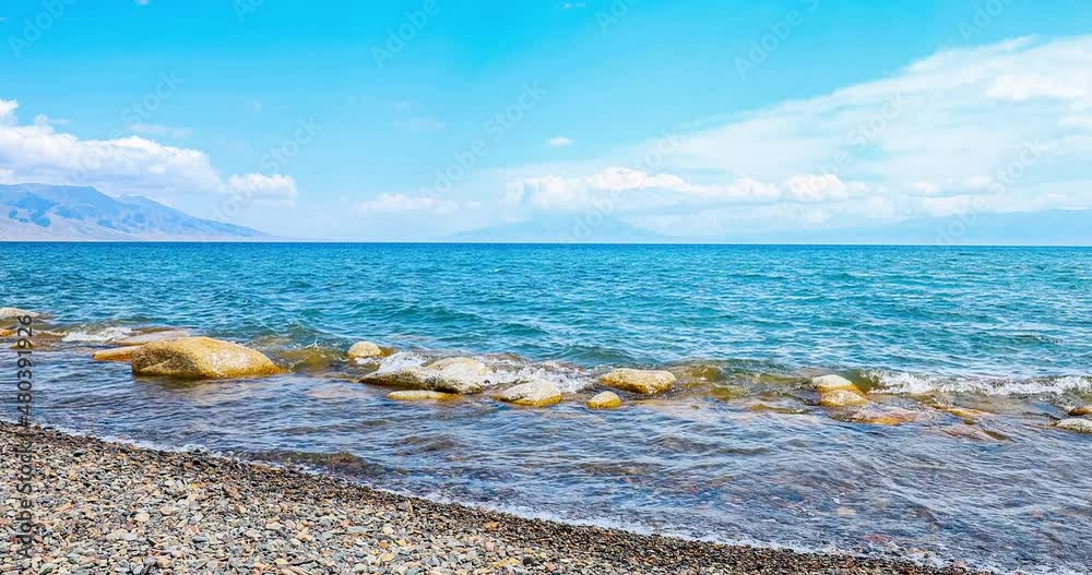 Sailimu Lake natural scenery in Xinjiang, China. Lake water lapping beach on shore. Clear lake water and beach with mountain under blue sky. Sailimu Lake natural scenery in Xinjiang, China. Lake water lapping beach on shore. Clear lake water and beach with mountain under blue sky.