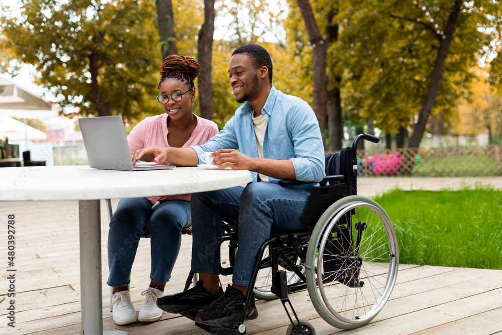 Cheerful impaired black man with female colleague using laptop together in outdoor cafe at autumn park Cheerful impaired black man with female colleague using laptop together in outdoor cafe at autumn park