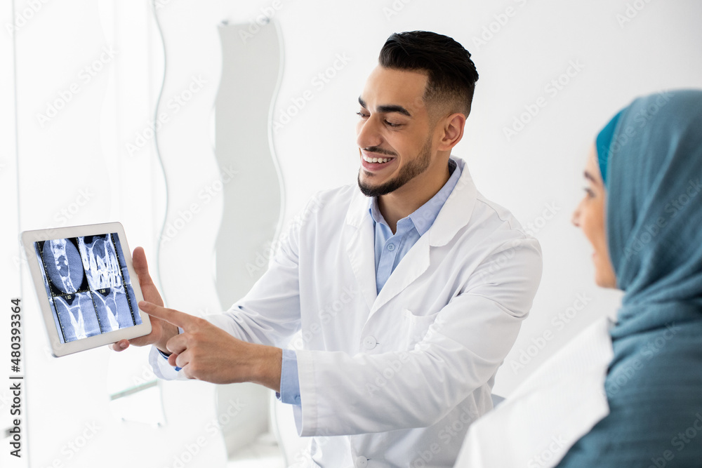 Arab Dentist Showing Teeth Xray On Digital Tablet To Muslim Female Patient