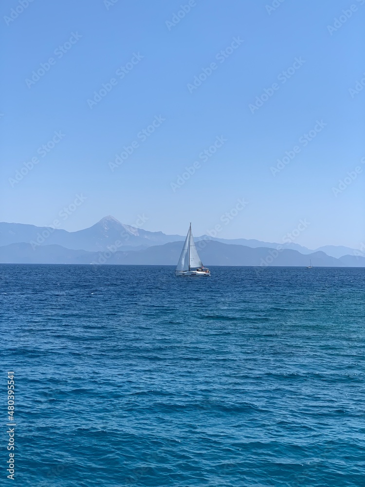 Naklejka premium Yacht with a white sail in the open sea. A sailboat sails in the ocean off the coast. Boat with a sail on the waves of a calm sea.