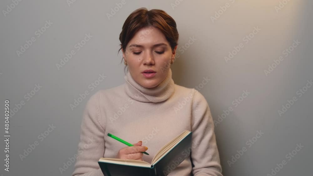 Short haired woman is speaking in front of camera while holding a book and pen on a gray studio wall Short haired woman is speaking in front of camera while holding a book and pen on a gray studio wall