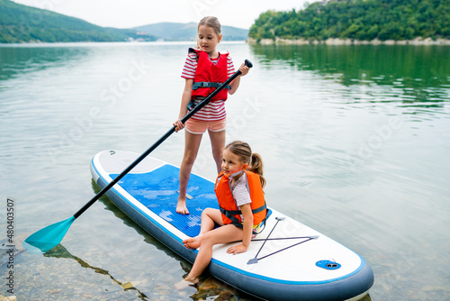 Girls padding on stand up paddle boarding on lake district. Children in swim life vest learning paddleboarding on SUP board. Family active leisure and local getaway with kids concept