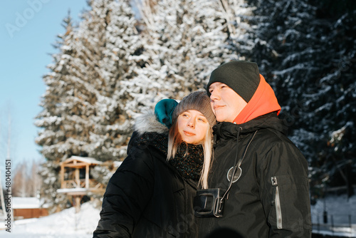 Heterosexual couple in love hugging on sunny winter day standing in snowy forest, outdoors. Young caucasian family enjoying nature together, looking away. Romantic leisure, valentines day concept